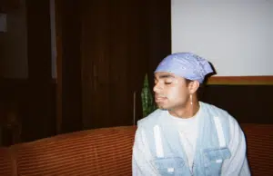 A person wearing a purple bandana and denim vest sits on an orange couch with closed eyes.